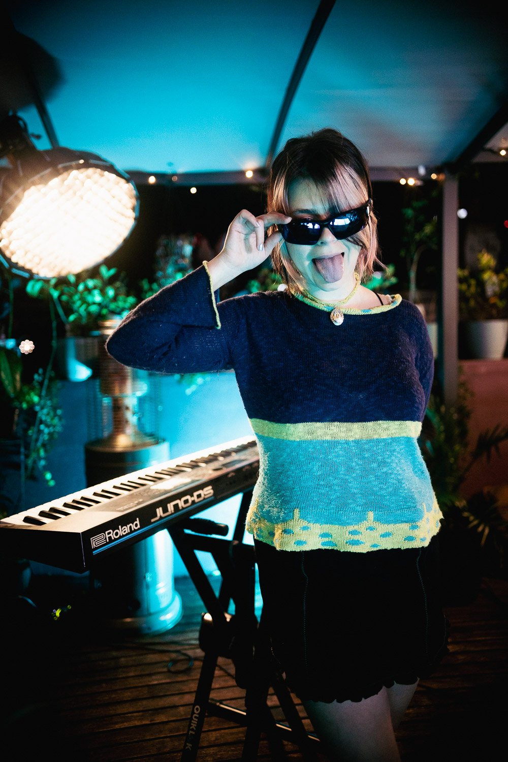 Lola Scott playfully adjusting sunglasses beside keyboard on rooftop stage.
