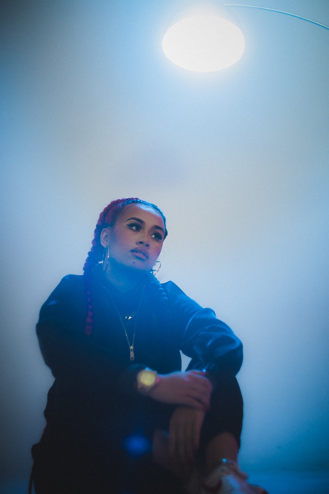 A.GIRL seated in studio lighting with braided hair, resting hand near chin.
