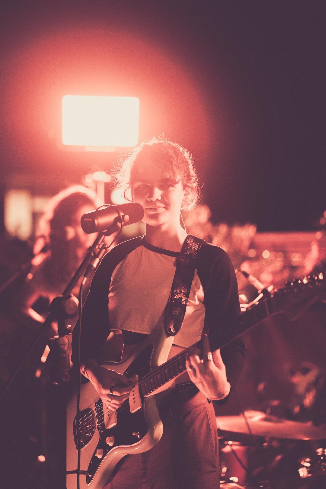 MAY-A singing into microphone while playing electric guitar under warm rooftop lighting.