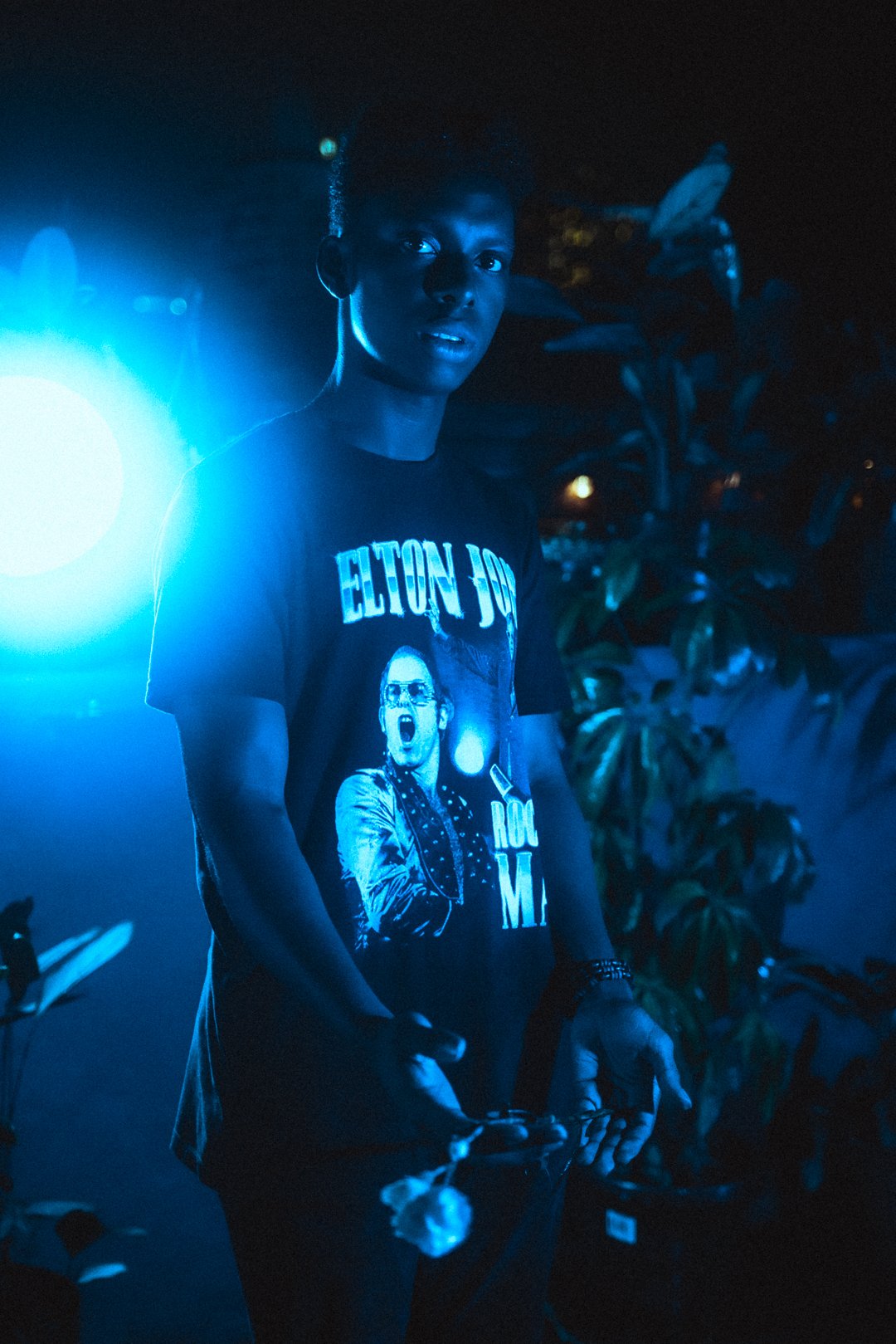 Stevan standing in blue light holding a small flower at rooftop stage.