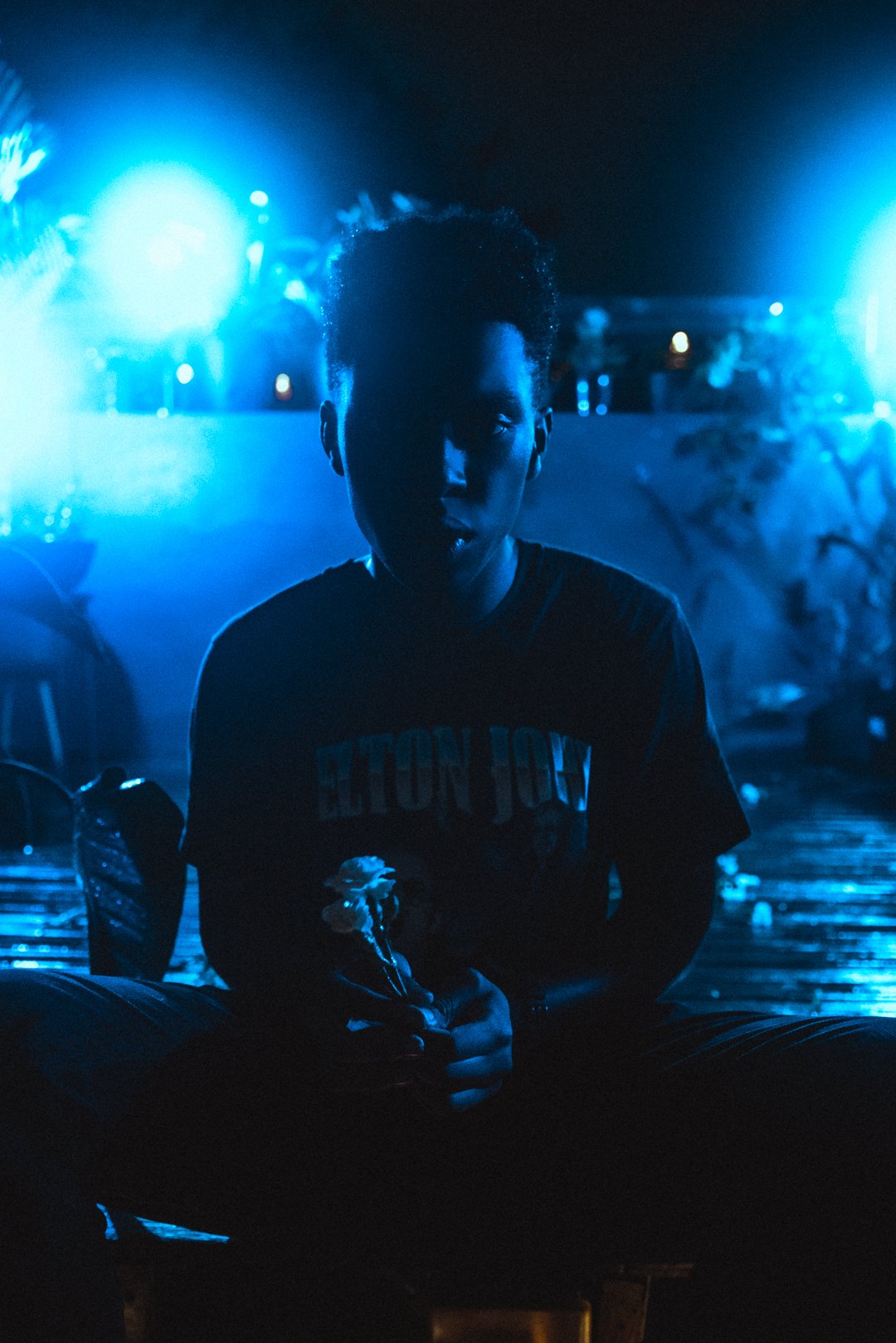 Stevan seated on rooftop holding flower under blue lights after rainfall.