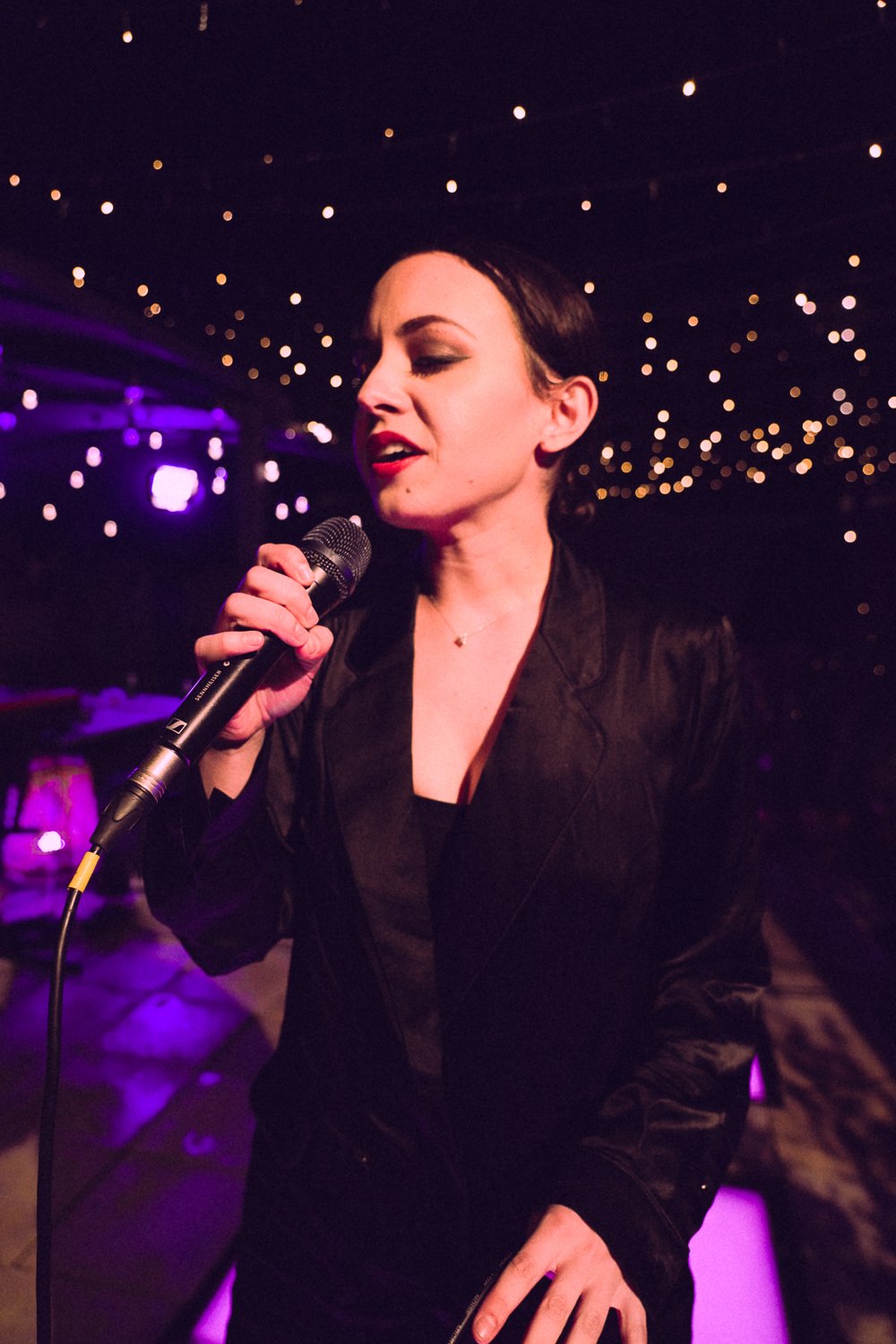 Sahara Beck singing in profile with microphone under purple rooftop lighting.