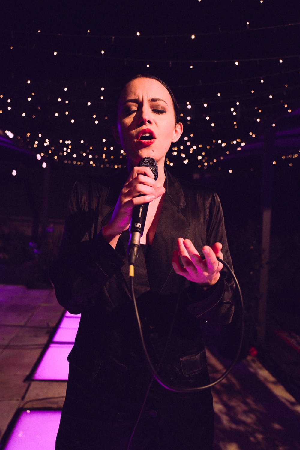 Sahara Beck sings passionately into a handheld microphone, eyes closed, gesturing beneath glowing rooftop string lights