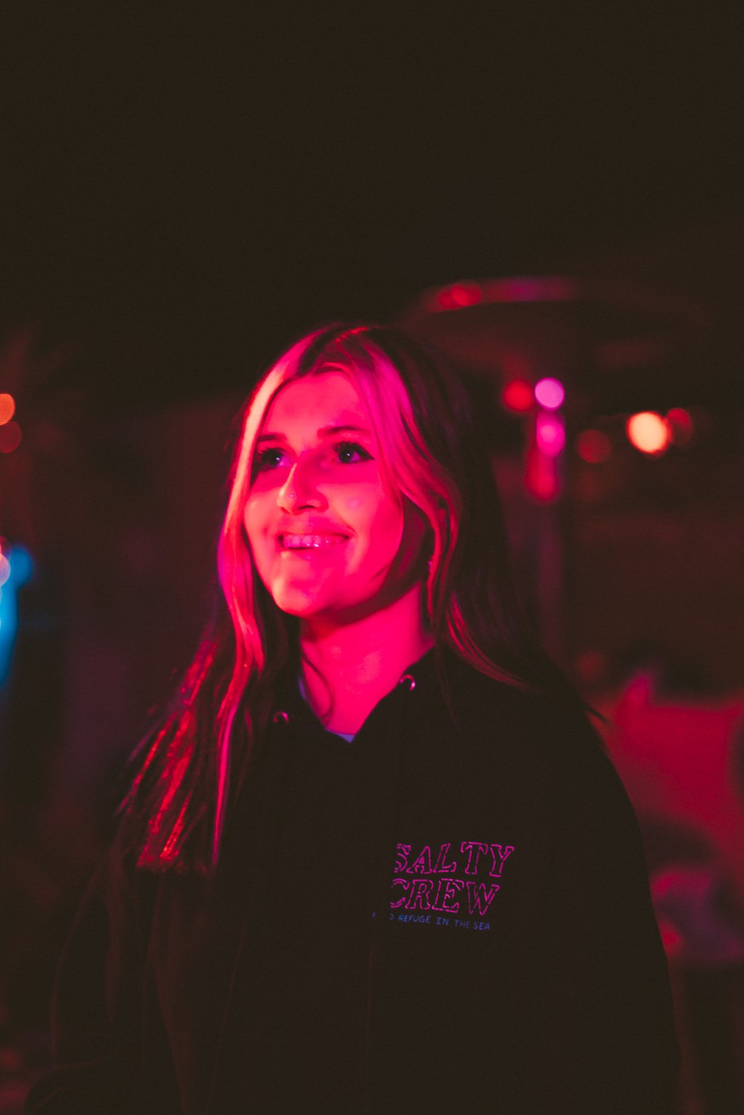 Abby Bella May smiling between songs under warm pink stage lighting.