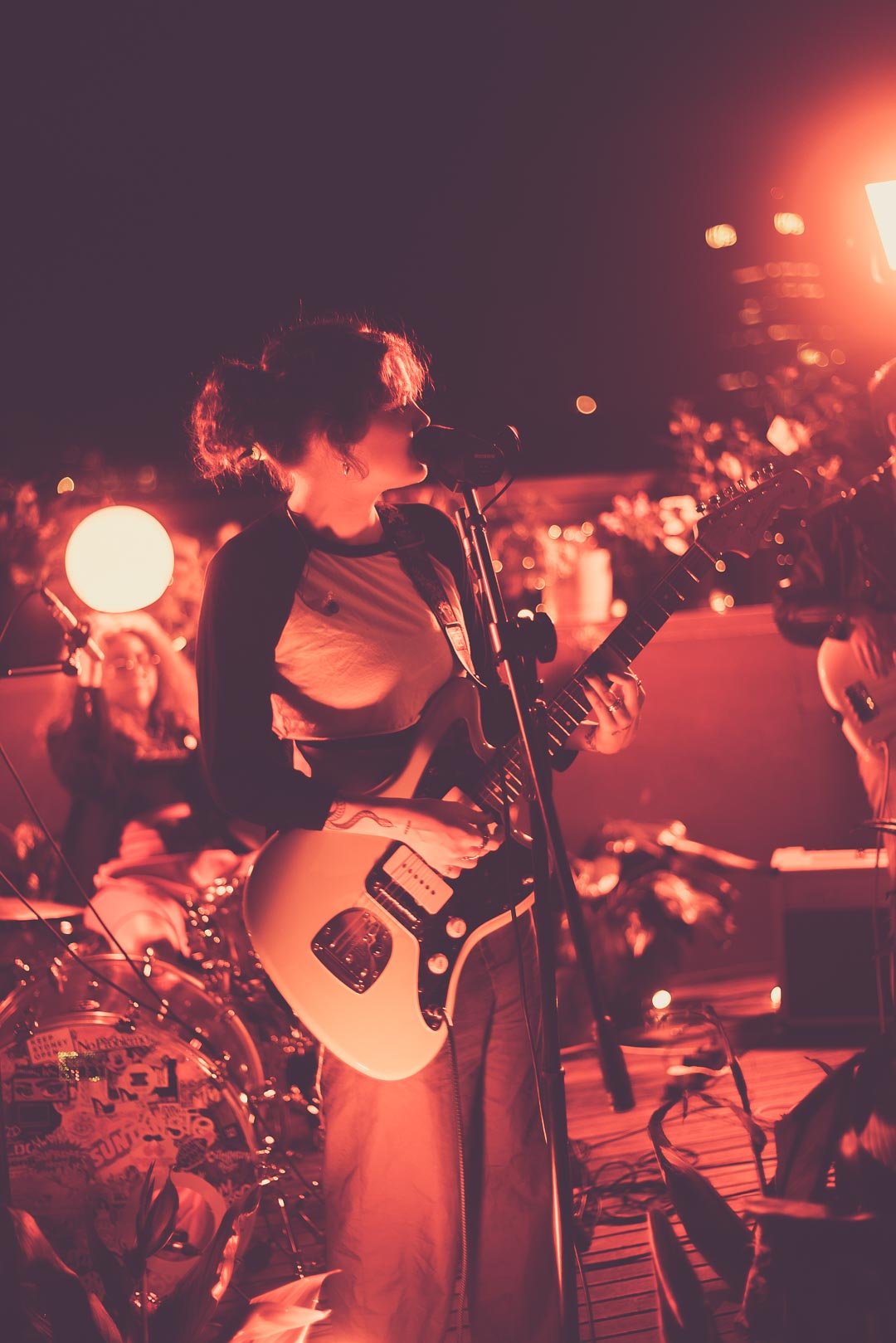 MAY-A playing electric guitar in profile at microphone under red stage lighting.